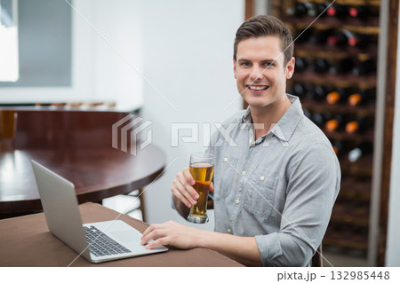 Man in late twenties typing on silver laptop holding beer glass at table in wine cellar 132985448
