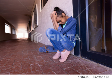 Elementary school girl sitting in school hallway hugging knees near door with backpack, copy space 132985458