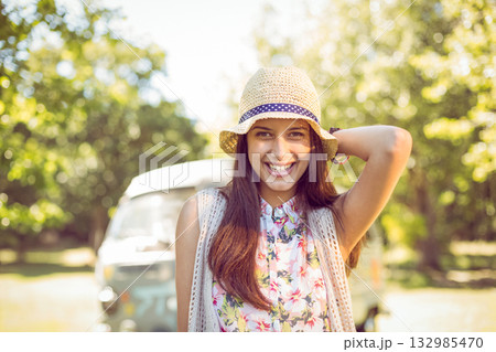 Woman standing in sunny park wearing floral blouse lifting hand behind straw hat near camper van Woman standing in sunny park wearing floral blouse lifting hand behind straw hat near camper van 132985470