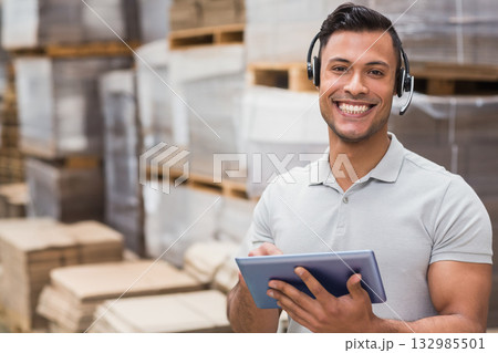 Adult male warehouse worker in grey polo with headset standing by boxes holding tablet, copy space 132985501