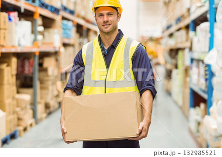 Male warehouse worker wearing hard hat and vest holding cardboard box in aisle among metal racks 132985521