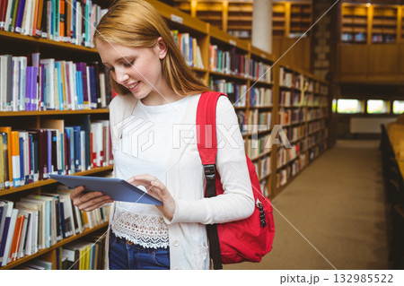 Female teenager standing in library aisle by bookshelves holding tablet and wearing red backpack Female teenager standing in library aisle by bookshelves holding tablet and wearing red backpack 132985522