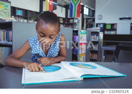 African American school-age girl leaning over open atlas in public library while pointing at map 132985524
