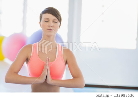 Woman wearing coral top standing with closed eyes pressing palms in yoga studio with exercise balls 132985536