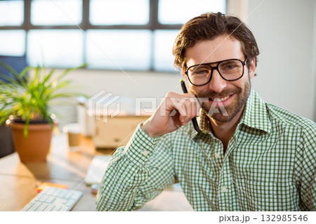Bearded man wearing green shirt sitting at desk in office holding pen near keyboard, potted plant 132985546