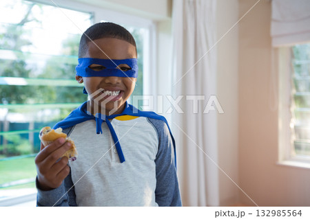 African American boy wearing blue mask and cape, holding sandwich near window at home, copy space African American boy wearing blue mask and cape, holding sandwich near window at home, copy space 132985564
