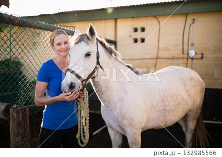 Woman holding braided lead rope touching white horse muzzle in stable yard by chain link fence Woman holding braided lead rope touching white horse muzzle in stable yard by chain link fence 132985566