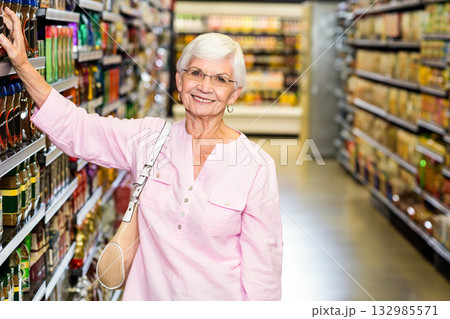 Senior woman browsing bottled goods in supermarket aisle wearing pink blouse and carrying beige bag 132985571