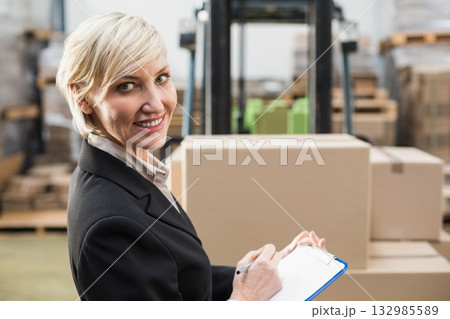 Middle-aged woman wearing blazer standing in warehouse holding clipboard near forklift, copy space Middle-aged woman wearing blazer standing in warehouse holding clipboard near forklift, copy space 132985589