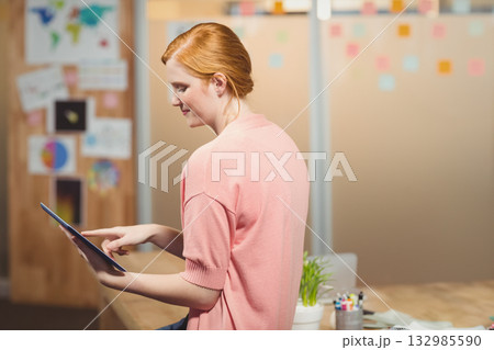 Woman sitting on edge of desk at office interacting with tablet computer beside potted plant Woman sitting on edge of desk at office interacting with tablet computer beside potted plant 132985590