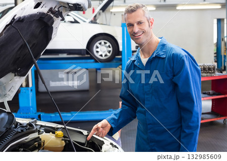 Male mechanic in blue coveralls pointing at coolant reservoir under hood in repair shop, copy space Male mechanic in blue coveralls pointing at coolant reservoir under hood in repair shop, copy space 132985609