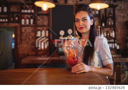 Woman in twenties holding orange-red cocktail with straw at bar counter with brick wall, copy space Woman in twenties holding orange-red cocktail with straw at bar counter with brick wall, copy space 132985626