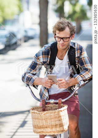 Man cycling with backpack and wicker basket along tree-lined sidewalk while checking smartphone 132985630