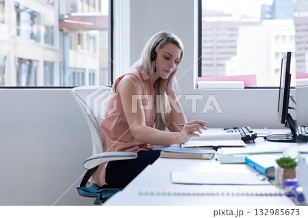 Fair-skinned woman wearing headset reading and marking notes at office desk with monitor, succulent 132985673