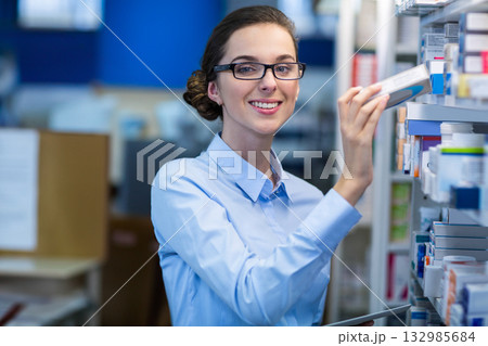 Teenage pharmacy assistant removing medicine box from metal shelf in pharmacy aisle with tablet 132985684