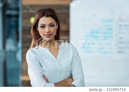 Female professional standing with arms crossed in office with whiteboard and glass partition 132985710