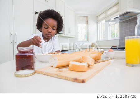 African American boy sitting at modern kitchen island eating cereal with spoon and orange juice 132985729