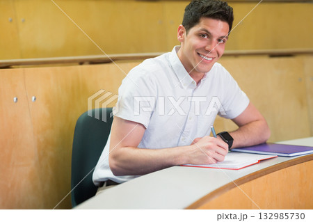 Male student writing with pen in notebook at university lecture hall curved desk wearing smartwatch 132985730