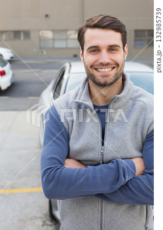 Male crossing arms and standing in parking lot in front of silver sedan near building windows 132985739