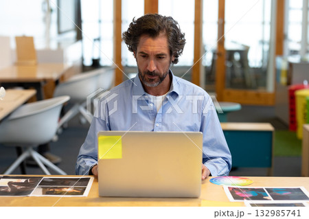 Mature adult man working on silver laptop at desk in creative office with color wheel chart Mature adult man working on silver laptop at desk in creative office with color wheel chart 132985741
