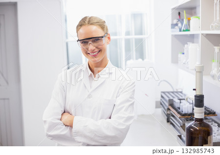 Female scientist smiling while standing in bright lab by bench with pipettes wearing white coat Female scientist smiling while standing in bright lab by bench with pipettes wearing white coat 132985743