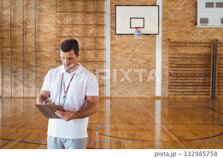 Mature adult male coach holding clipboard with pen near basketball hoop and wall bars in gymnasium 132985758