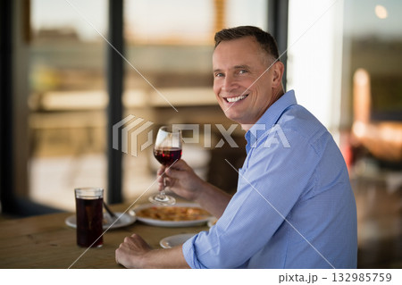 Middle-aged man holding red wine glass, smiling at wooden restaurant table with pasta and dark soda Middle-aged man holding red wine glass, smiling at wooden restaurant table with pasta and dark soda 132985759