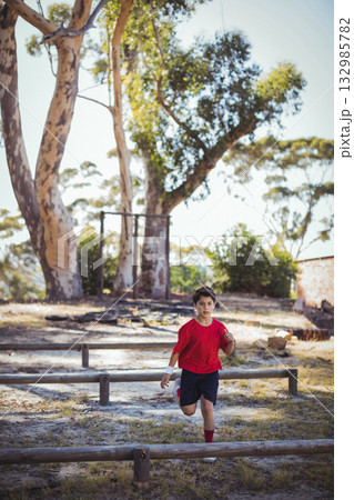 Boy running along dirt clearing navigating wooden-beam obstacle course holding oval ball in park 132985782