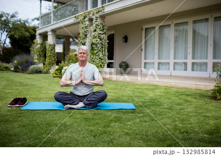 White senior man practicing yoga on blue mat on backyard lawn with columns wrapped in vines 132985814