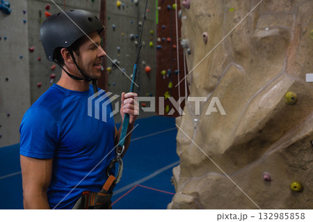 Mid adult man standing at rock-climbing gym wearing helmet and harness holding belay rope Mid adult man standing at rock-climbing gym wearing helmet and harness holding belay rope 132985858
