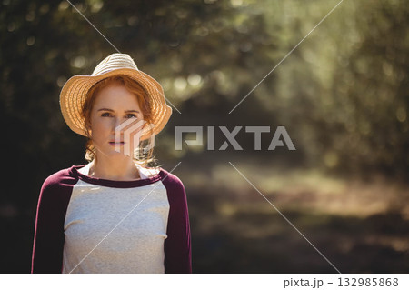 Woman standing outdoors wearing straw sun hat and raglan shirt on sunlit woodland path, copy space 132985868