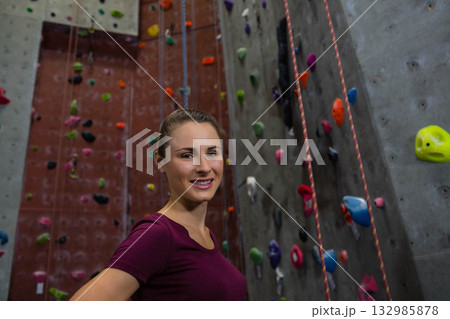 Woman in twenties wearing burgundy tee smiling against climbing wall with multicolored holds, ropes 132985878