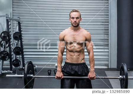 Shirtless man wearing black shorts holding loaded barbell at thigh level at gym by squat rack Shirtless man wearing black shorts holding loaded barbell at thigh level at gym by squat rack 132985890