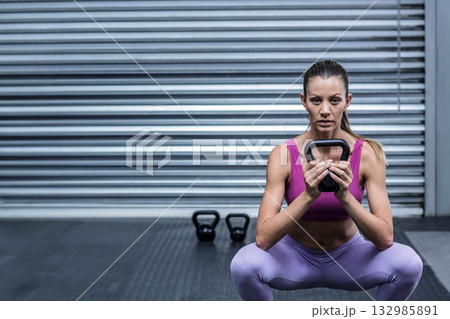 Woman in fuchsia bra, leggings squatting, gripping black kettlebell under metal shutter at gym 132985891
