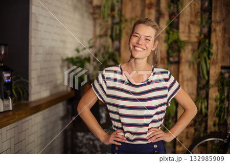 Smiling woman standing at coffee shop counter with coffee grinder wooden wall and bicycle wheel 132985909