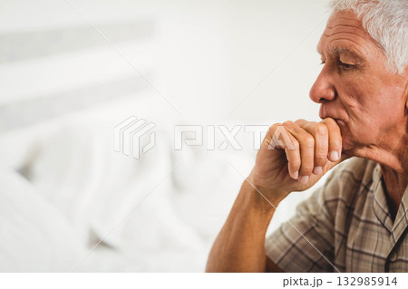 Senior man sitting on bed in bedroom resting chin on clasped hand beside headboard, copy space Senior man sitting on bed in bedroom resting chin on clasped hand beside headboard, copy space 132985914