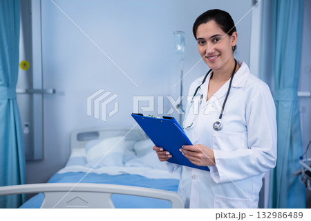 Smiling Indian doctor holding clipboard, wearing stethoscope at bedside with IV pole, copy space Smiling Indian doctor holding clipboard, wearing stethoscope at bedside with IV pole, copy space 132986489