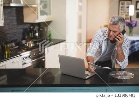 Senior man standing at kitchen island talking on smartphone, reading papers by laptop, copy space 132986493