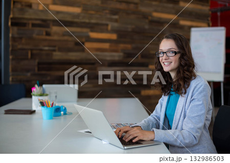 Woman in teal blouse, glasses typing on laptop at coworking table with notebook, copy space 132986503