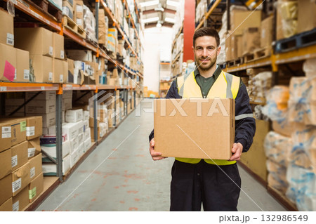 Male warehouse worker wearing safety vest holding cardboard box in aisle with metal shelving Male warehouse worker wearing safety vest holding cardboard box in aisle with metal shelving 132986549
