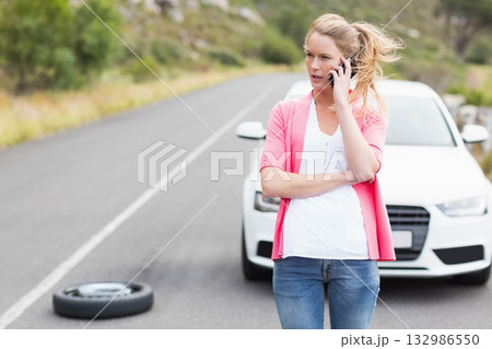 Woman standing on rural paved road beside white sedan holding smartphone and spare tire, copy space Woman standing on rural paved road beside white sedan holding smartphone and spare tire, copy space 132986550