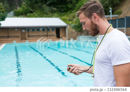 Male coach holding stopwatch and checking display standing poolside with lane ropes, copy space Male coach holding stopwatch and checking display standing poolside with lane ropes, copy space 132986568