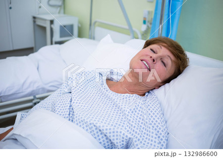 Senior woman lying reclined on white pillows in hospital bed with hospital gown and medical monitor 132986600