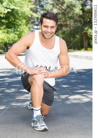 Male runner kneeling on park road holding knee smiling in sportswear with running shoes, bib 1604 Male runner kneeling on park road holding knee smiling in sportswear with running shoes, bib 1604 132986611