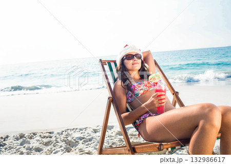 Woman in holiday hat reclining on wooden chair at water's edge holding pink cocktail, copy space Woman in holiday hat reclining on wooden chair at water's edge holding pink cocktail, copy space 132986617