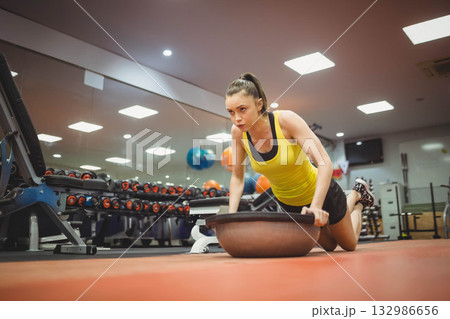 Woman wearing yellow tank top and black shorts performing push-up kneeling on BOSU ball at gym 132986656