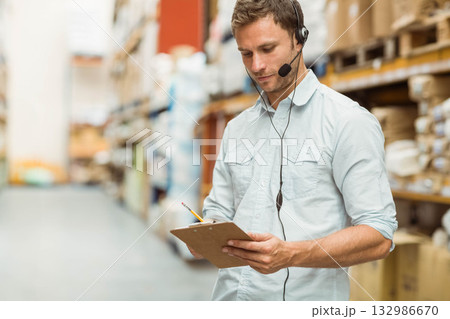 Mid-adult man checking inventory on clipboard in warehouse aisle with headset and boxes, copy space Mid-adult man checking inventory on clipboard in warehouse aisle with headset and boxes, copy space 132986670