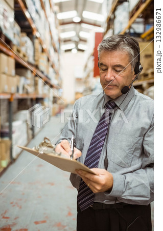 Man supervising inventory in warehouse aisle wearing tie, holding clipboard and headset, copy space Man supervising inventory in warehouse aisle wearing tie, holding clipboard and headset, copy space 132986671