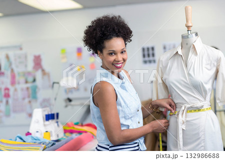 African American woman fitting dress on mannequin in studio with sewing machine and tape measure 132986688