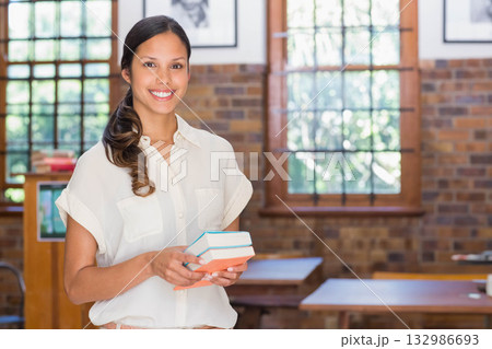Woman librarian smiling and holding stack of textbooks in classroom with wooden lectern Woman librarian smiling and holding stack of textbooks in classroom with wooden lectern 132986693